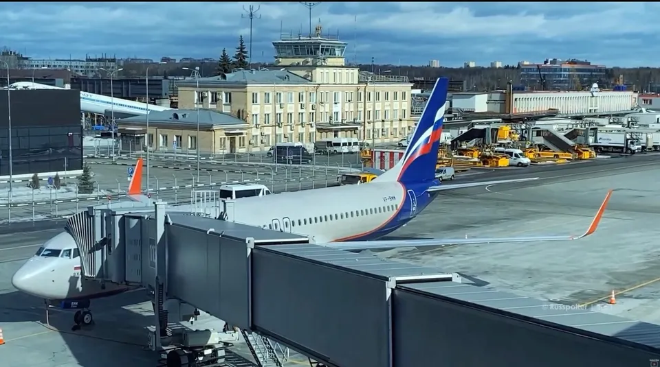 Un avion alb cu coadă albastră și roșie stă pe pista unui aeroport lângă o punte de îmbarcare, având clădiri și vehicule în fundal.