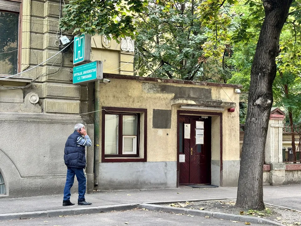 Un bărbat cu vestă albastră și pantaloni de denim stă pe trotuar lângă o clădire cu o fereastră și o ușă maro.