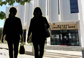 Două silhuete feminine merg în fața unei clădiri albe, cu trepte, intrare vitrată și inscripție deasupra ușii.