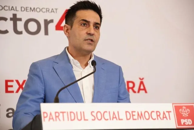 Un bărbat cu sacou albastru și cămașă albă vorbește la un microfon aflat în fața unui panou cu text colorat.