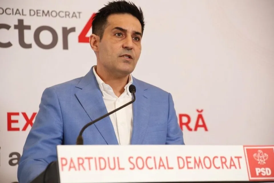 Un bărbat cu părul scurt și sacou albastru stă în spatele unui podium alb cu inscripții roșii și vorbește la un microfon.