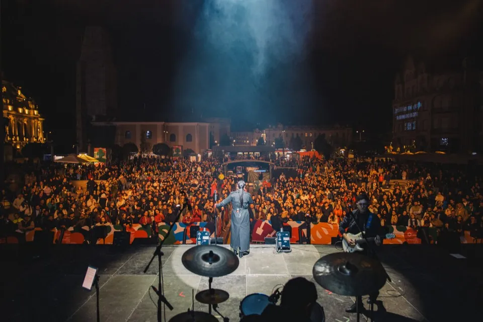 O mulțime de oameni stă în fața unei scene pe care se află un cântăreț și instrumente muzicale sub lumini de spectacol.