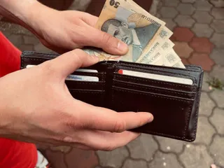 Două mâini țin un portofel deschis de culoare închisă care conține bancnote și carduri, în timp ce degetele de sus țin câteva bancnote.
