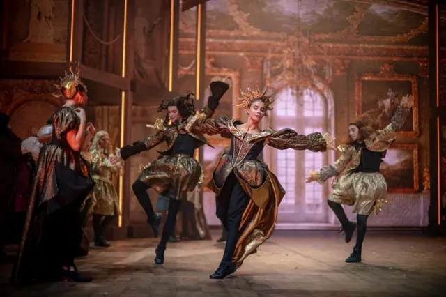 Patru persoane în costume de scenă aurii și negre execută mișcări de dans pe o scenă de teatru iluminată.