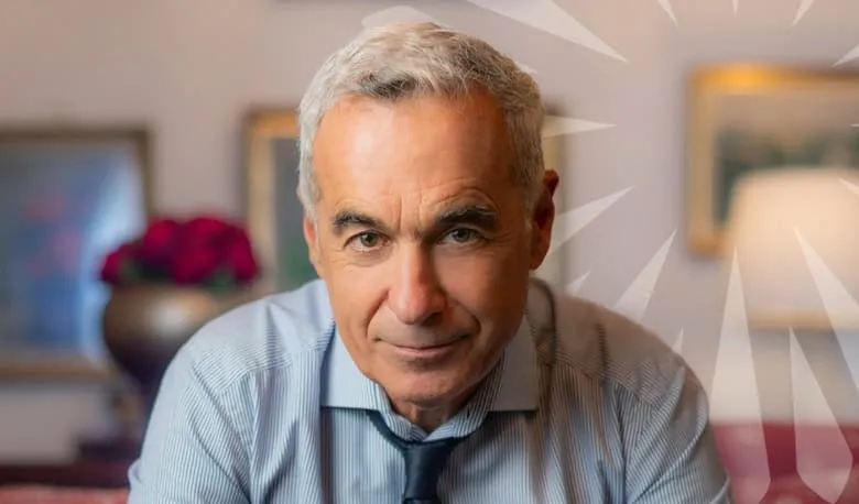 Un bărbat cu părul cărunt și cămașă albastră deschisă privește spre cameră într-un interior cu tablouri și o lampă în fundal.