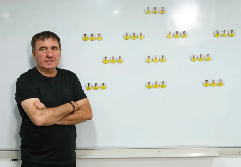 Un bărbat cu tricou negru stă cu brațele încrucișate în fața unui panou alb pe care sunt lipite mai multe imagini mici cu fețe de oameni.