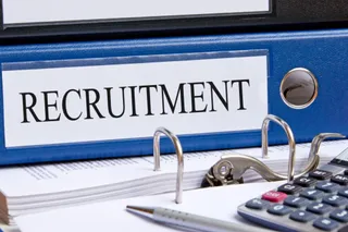 Un dosar albastru cu eticheta RECRUITMENT stă pe un teanc de hârtii lângă un calculator și un pix.