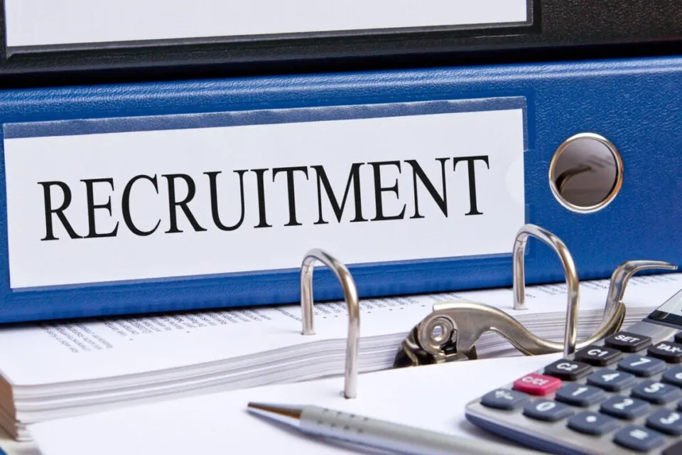 Un dosar albastru cu eticheta RECRUITMENT stă pe un teanc de hârtii lângă un calculator și un pix.