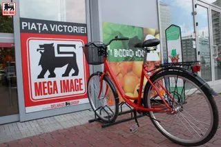 O bicicletă roșie cu un coș negru este parcată în fața unui magazin care prezintă un afiș cu un leu și textul Mega Image.