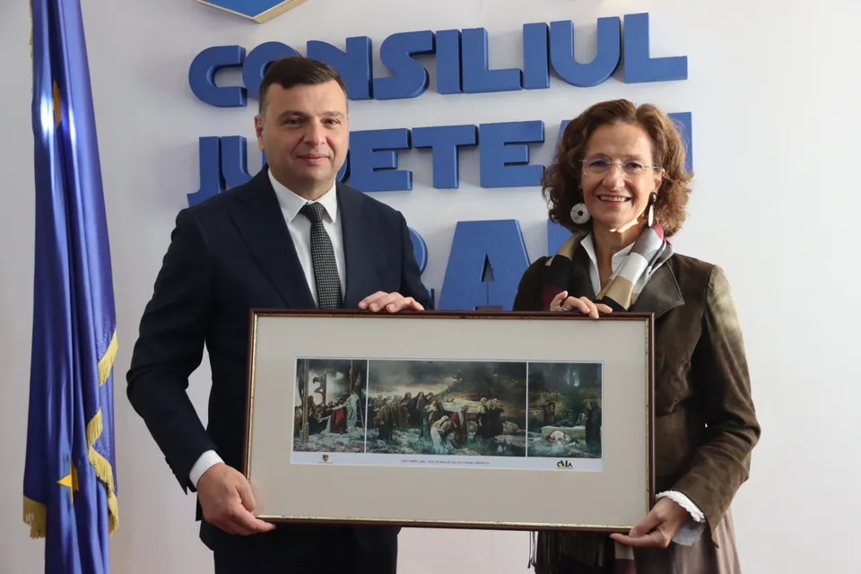 Un bărbat în costum și o femeie în sacou țin împreună un tablou încadrat în fața unui perete cu litere albastre mari.
