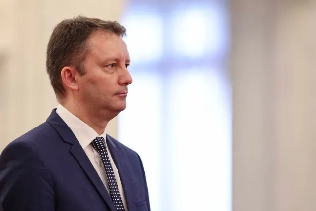 Un bărbat cu părul scurt, purtând sacou albastru, cămașă albă și cravată cu puncte, stă în profil într-un decor cu fundal deschis și neclar.