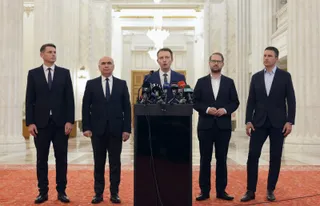 Cinci bărbați în costume scurte stau în picioare în fața unui podium cu microfoane într-o încăpere cu coloane albe și podea cu covor roșu.