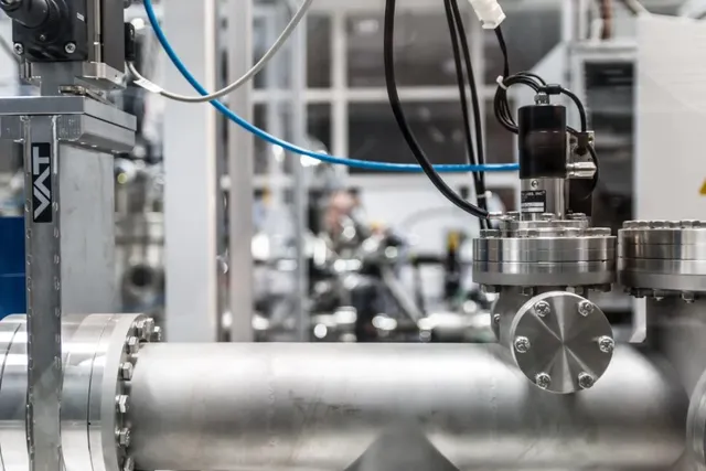 O conductă metalică de mari dimensiuni este conectată la diverse componente industriale și cabluri într-un mediu de laborator sau fabrică.