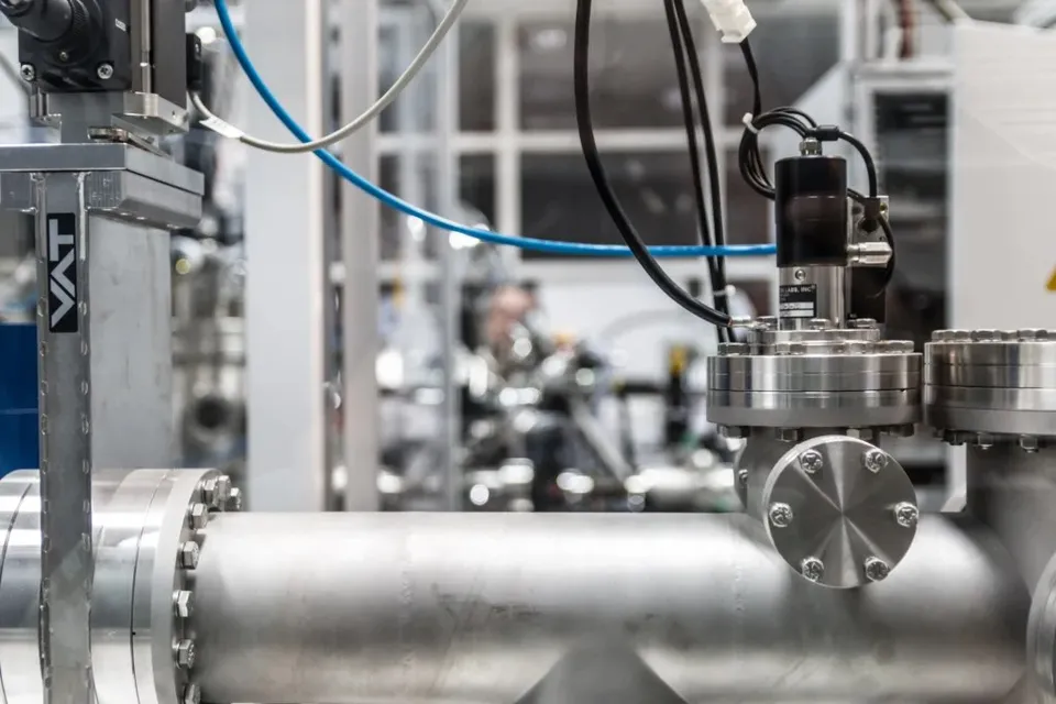 O conductă metalică de mari dimensiuni este conectată la diverse componente industriale și cabluri într-un mediu de laborator sau fabrică.