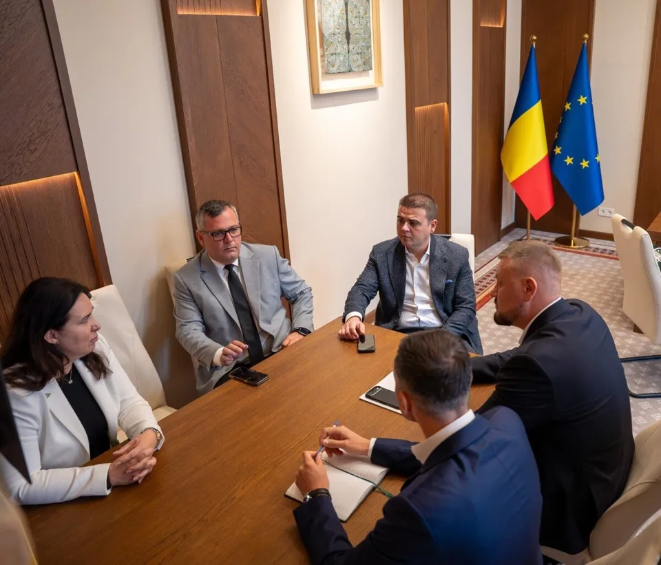 Cinci persoane stau la o masă de lemn într-o încăpere cu panouri de lemn și două steaguri în fundal.