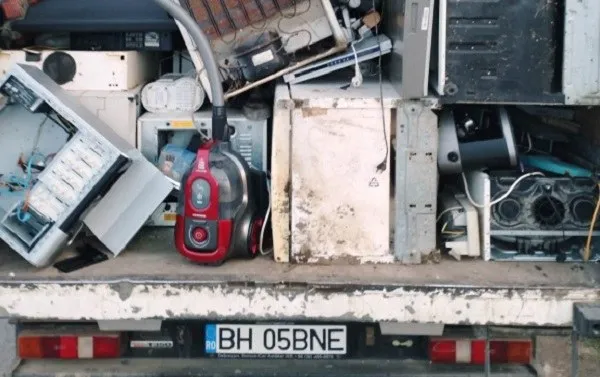 Diverse echipamente electrice și electronice uzate, inclusiv un aspirator roșu și unități de calcul, sunt depozitate pe platforma unui vehicul cu număr de înmatriculare BH 05BNE.