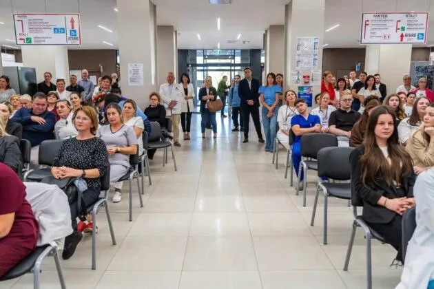 Oamenii stau așezați pe scaune negre dispuse în rânduri pe un hol cu podea lucioasă, privind spre centrul încăperii.