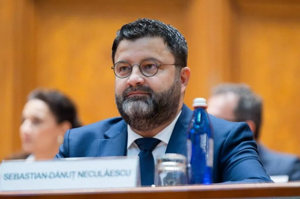 Un bărbat cu barbă și ochelari, purtând sacou albastru și cravată, stă la o masă lângă o sticlă de apă și un pahar.