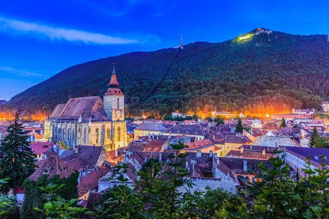 O vedere panoramică asupra unui oraș cu clădiri cu acoperișuri înclinate, situat la poalele unui munte copacos sub un cer albastru.