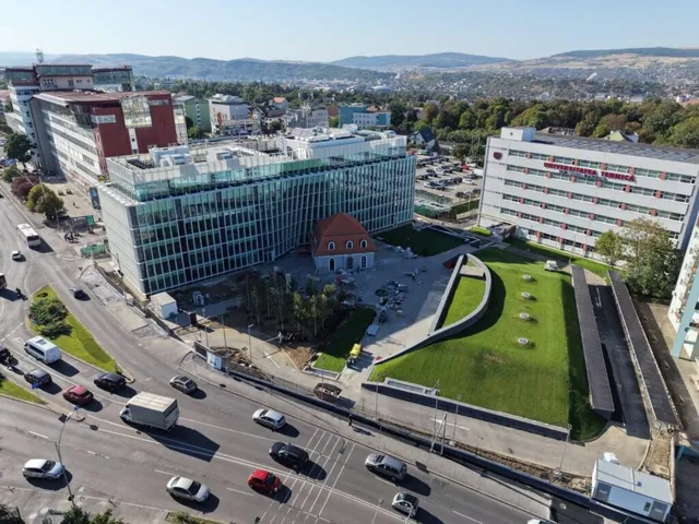 O vedere aeriană prezintă clădiri moderne de birouri, o stradă cu mașini și camioane, un teren de fotbal și dealuri în fundal.