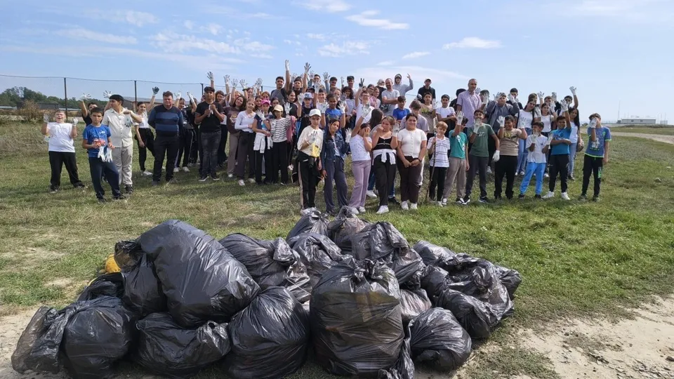 Un grup numeros de copii și adulți stă pe iarbă, în spatele unor saci negri voluminoși, sub cer deschis.