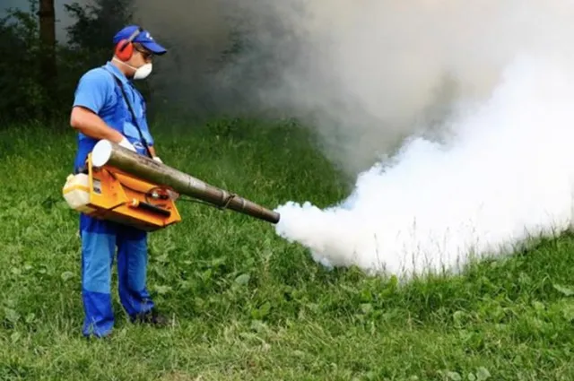 Un bărbat cu echipament de protecție și mască poartă un aparat de pulverizare care scoate un nor de fum alb peste iarbă.