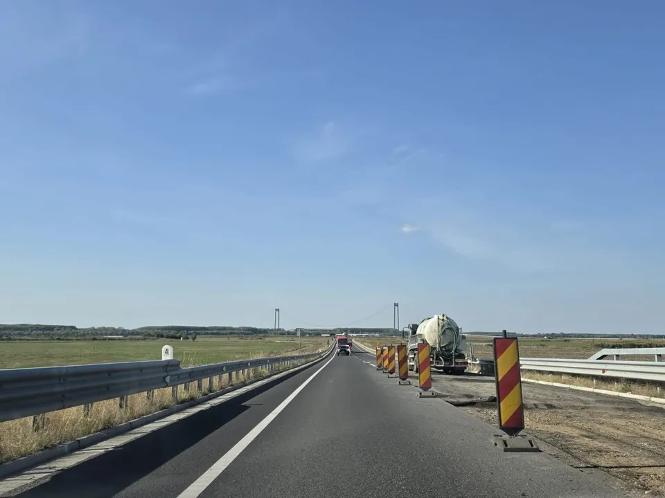 O șosea dreaptă cu barieră metalică și marcaje, delimitată de câmpuri, are indicatoare portabile și un utilaj pe banda din dreapta, sub cer senin.
