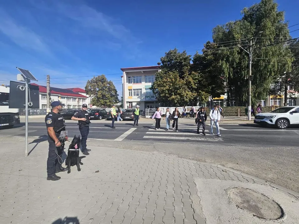 Un polițist cu un câine negru stă pe trotuar, în timp ce alți polițiști și persoane merg pe strada din fața unei clădiri albe.