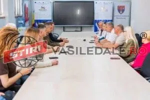 O grupă de persoane stă la o masă lungă într-o încăpere cu un ecran negru montat pe perete în fundal.