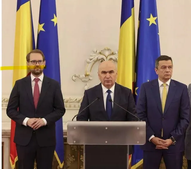 Trei bărbați în costume și cravate stau în picioare în fața unor steaguri, în timp ce cel din mijloc se află în spatele unui podium.