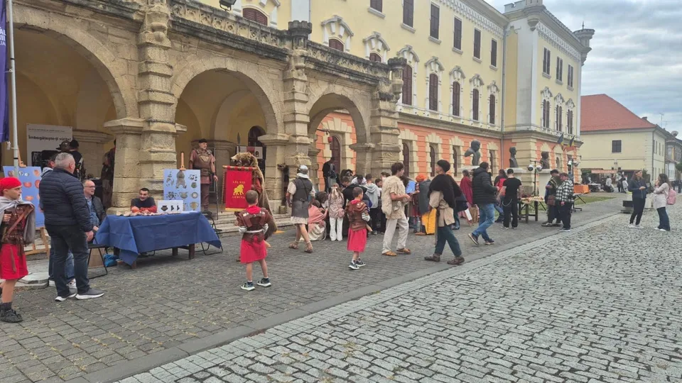 Oamenii stau pe o stradă pavată în fața unei clădiri mari cu arcade, în timp ce unii poartă costume istorice.