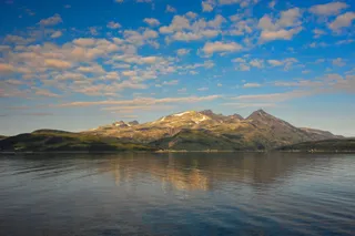 Munți cu zone de zăpadă sunt reflectați în apa calmă sub un cer albastru cu nori albi.