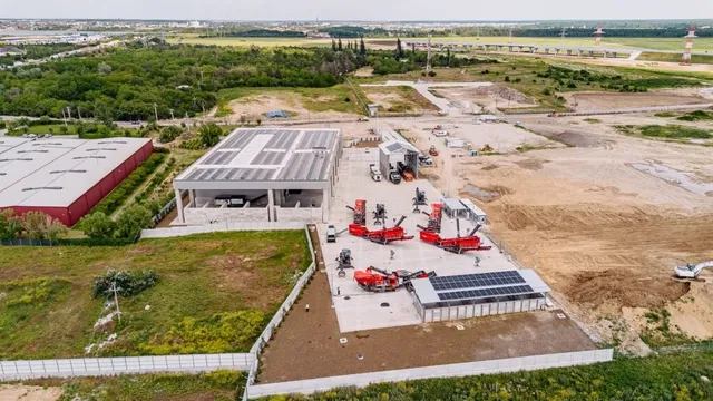O vedere aeriană prezintă o clădire industrială cu panouri solare pe acoperiș, utilaje de construcții roșii și o zonă de depozitare cu pământ.
