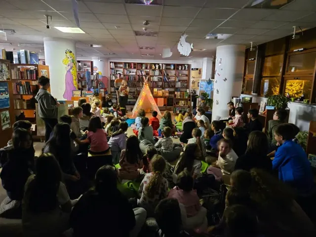 Un grup de copii și adulți stau pe podeaua unei biblioteci în fața unui cort textil de culoare portocalie, înconjurată de rafturi pline de cărți.