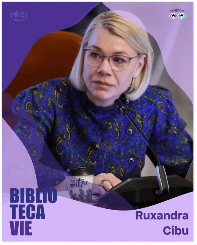 O femeie cu ochelari și părul blond, purtând o bluză albastră cu model paisley, stă la o masă lângă un microfon și un pahar de cafea.