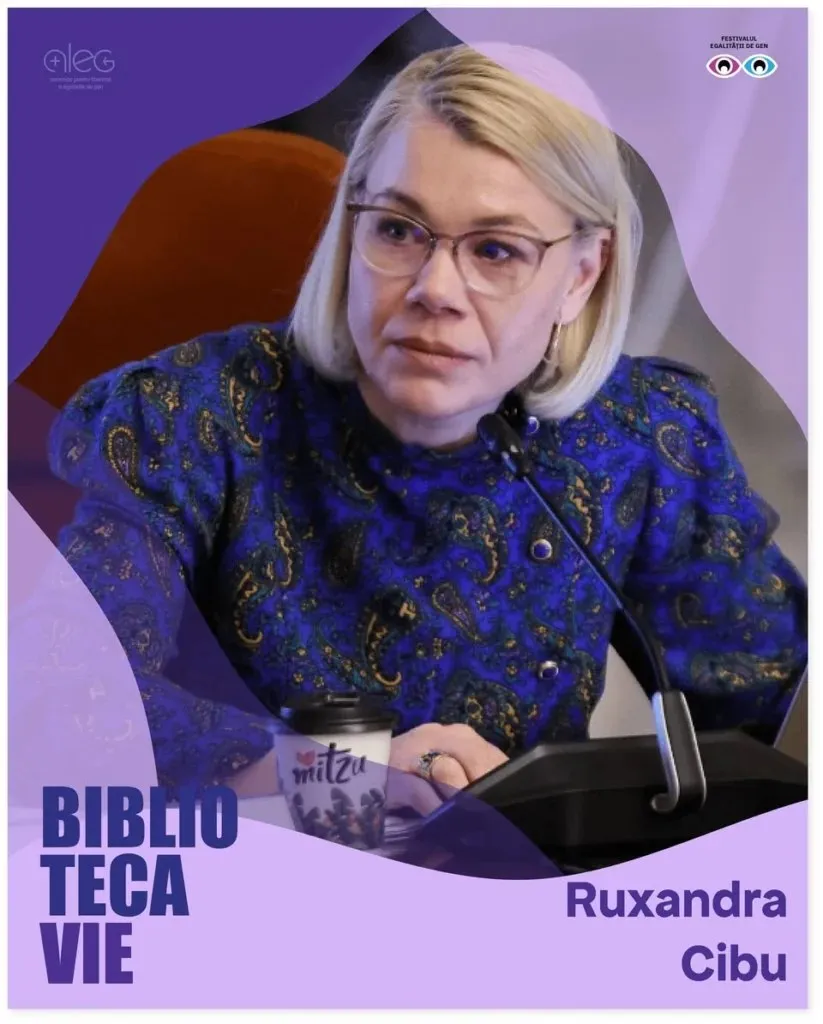 O femeie cu ochelari și părul blond, purtând o bluză albastră cu model paisley, stă la o masă lângă un microfon și un pahar de cafea.