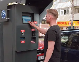 Un bărbat cu tricou negru apasă pe un ecran tactil al unui aparat de plată de parcare situat pe o stradă cu mașini parcate.