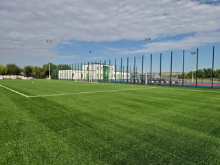 Un teren de fotbal cu iarbă artificială verde este delimitat de linii albe, având un clădire albă și stâlpi de iluminat în fundal.