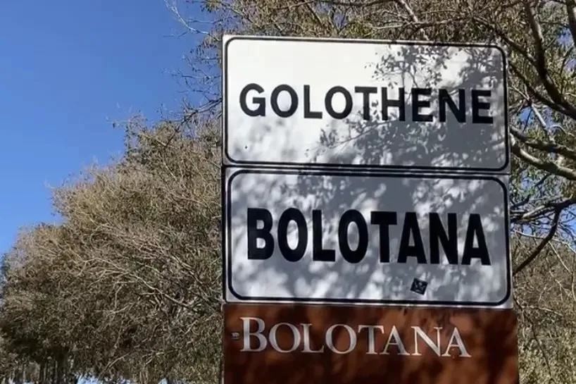 Două semne rutiere albe cu text negru indică locațiile Golothene și Bolotana sub un cer albastru și ramuri de copaci.