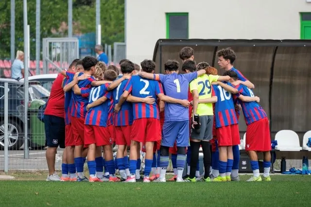 Un grup de tineri sportivi în echipamente de fotbal albastre și roșii stau într-un cerc cu brațele pe umerii colegilor pe un teren verde.