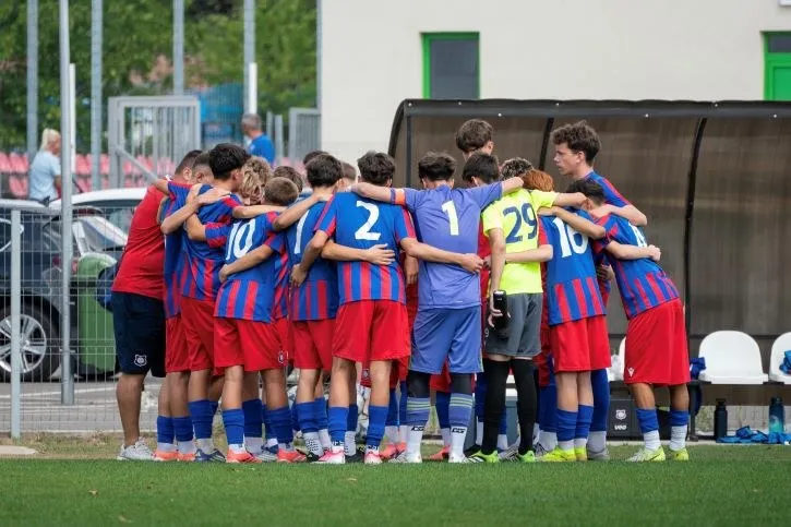 Un grup de tineri sportivi în echipamente de fotbal albastre și roșii stau într-un cerc cu brațele pe umerii colegilor pe un teren verde.