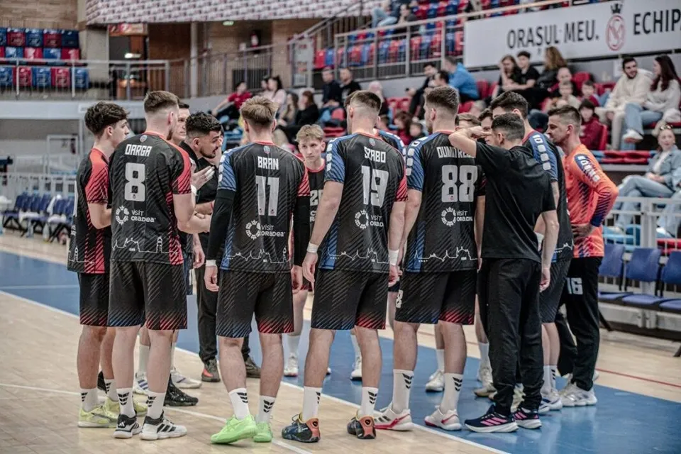 Un grup de sportivi în echipamente de handbal negre și roșii stau adunați într-o sală de sport cu tribunele ocupate în fundal.