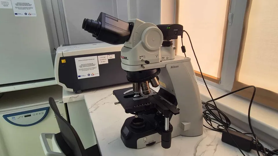 Un microscop de laborator de culoare albă este poziționat pe o masă de lucru lângă un aparat medical și un cablu de alimentare negru.