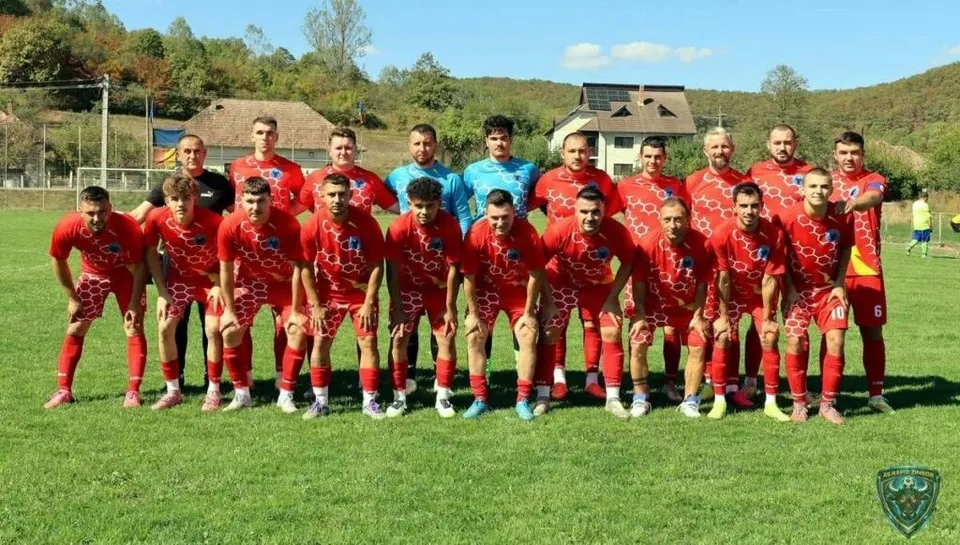 O echipă de fotbaliști îmbrăcați în echipamente roșii și albe stau în rânduri pe un teren de fotbal verde sub un cer senin.
