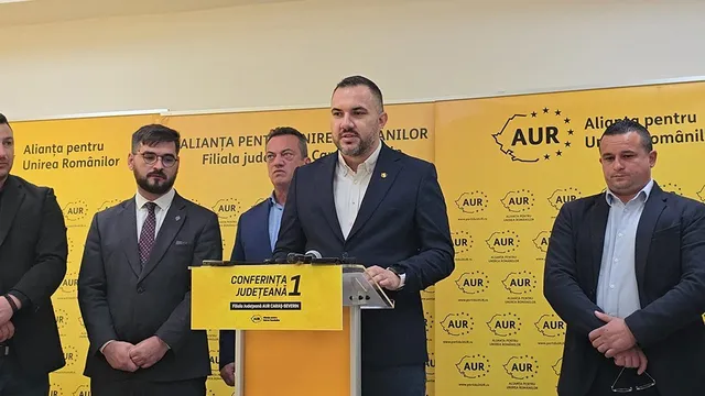 Cinci bărbați în costume și cămași stau în spatele unui podium galben pe un fundal cu logouri AUR repetate.