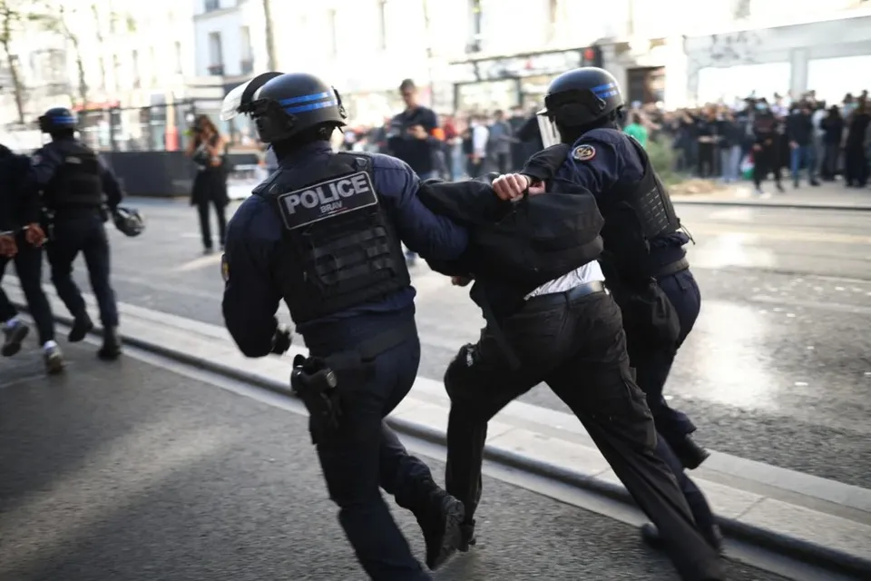 Trei polițiști în echipament tactice și casci întđen restricționează mișcările unei persoane îmbrăcate în negru pe o stradă urbană.