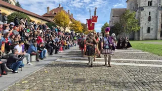 Oamenii în costume istorice merg pe un drum pavat în fața unui grup de spectatori așezați pe o bancă lângă clădiri vechi.
