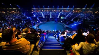 O mulțime de oameni stă așezată în tribunele unui auditorium întunecat, privind spre o arenă circulară iluminată cu lumini albastre.