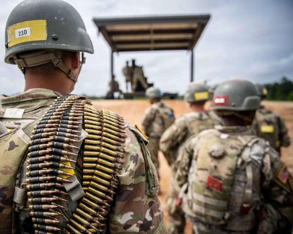 Soldați în uniforme de camuflaj și căști metalice stau cu spatele, purtând bandoliere cu gloanțe de calibru mare pe umeri.
