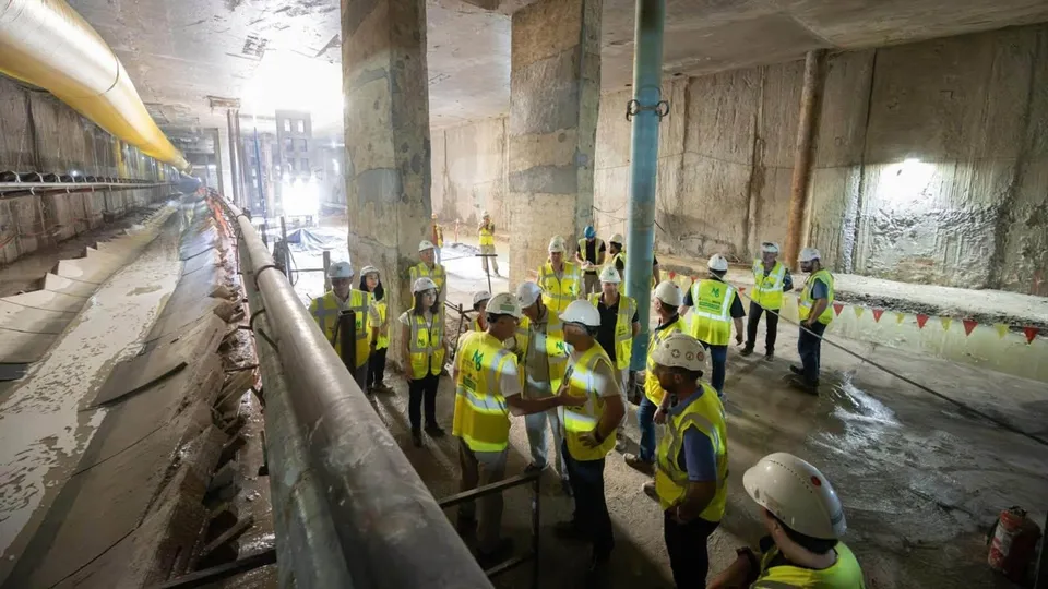 O grupă de muncitori cu căști și veste reflectorizante stă în interiorul unui șantier subteran lângă conducte și pereți de beton.
